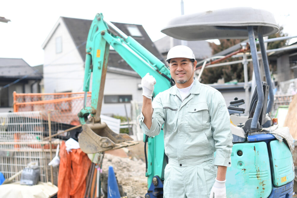 工事現場の重機の前でガッツポーズをする作業員の男性（カメラ目線）